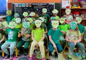 Dzieci siedzą w grupie na krzesłach. Mają bluzki w zielonych kolorach. Na twarzach maski ufoludków. Każdy w dłoniach trzyma instrument perkusyjny.